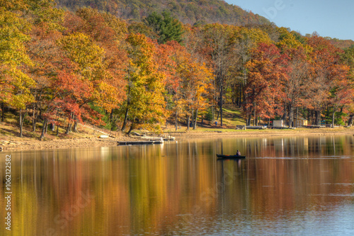 A serene lake scene captures vivid autumn colors as the trees reflect on the calm water. A lone boat drifts gently, enhancing the tranquil atmosphere.