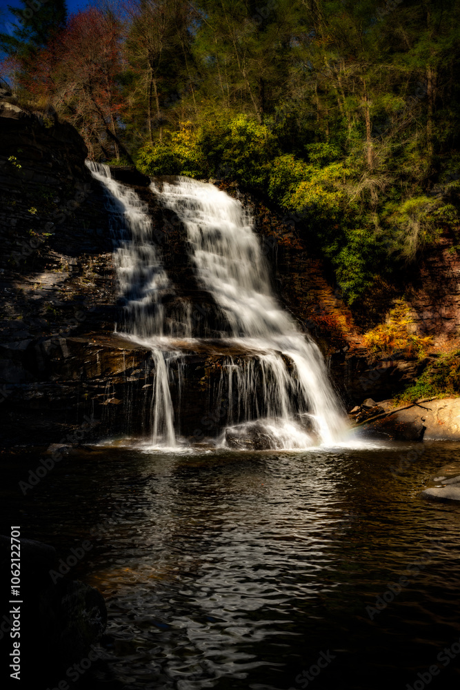 Obraz premium Water cascades down a rocky cliff into a tranquil pool, surrounded by lush greenery and trees. Sunlight highlights the vibrant colors of the foliage, creating a serene and natural scene.