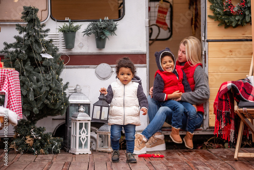 Little girl holding lantern stands outside decorated camper, with diversity family enjoying the holiday season together. Warm and cozy seasonal decor surrounds them. photo