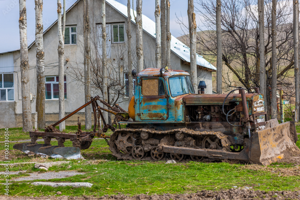 An old, rusty bulldozer with a plow Against two-story house surrounded by row of tall trees in a tranquil rural setting at spring day An old, rusty bulldozer with a plow Against two-story house surrounded by row of tall trees in a tranquil rural setting at spring day