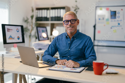 Smiling senior businessman sitting at his desk in a modern office, analyzing charts and working on a laptop, demonstrating confidence and expertise