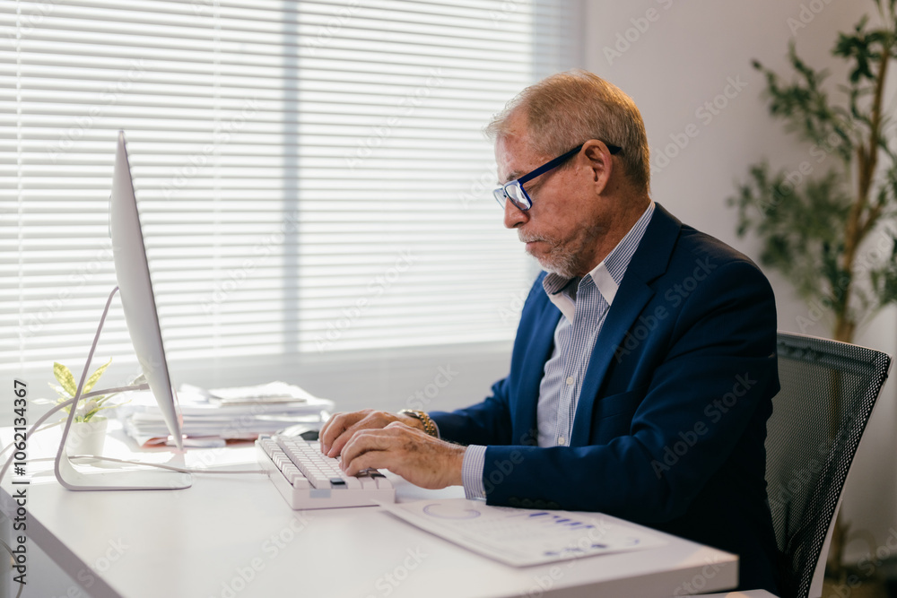 Focused mature manager typing on keyboard, working with computer and documents in bright office, demonstrating professionalism and expertise in business environment