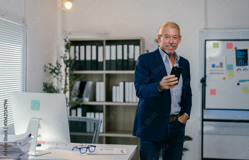 Smiling senior businessman using smartphone in modern office with hand in pocket, standing near desk with computer and documents, shelves with folders and whiteboard with graphs Smiling senior businessman using smartphone in modern office with hand in pocket, standing near desk with computer and documents, shelves with folders and whiteboard with graphs