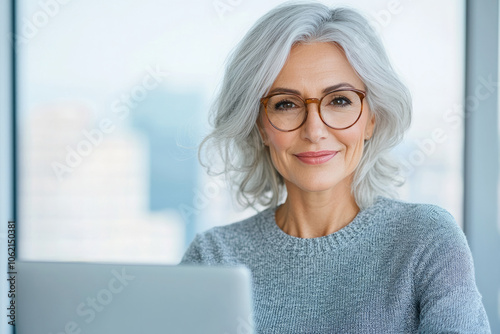 Free Download Smiling mature woman with glasses working on a laptop in a modern office - GFXLoad