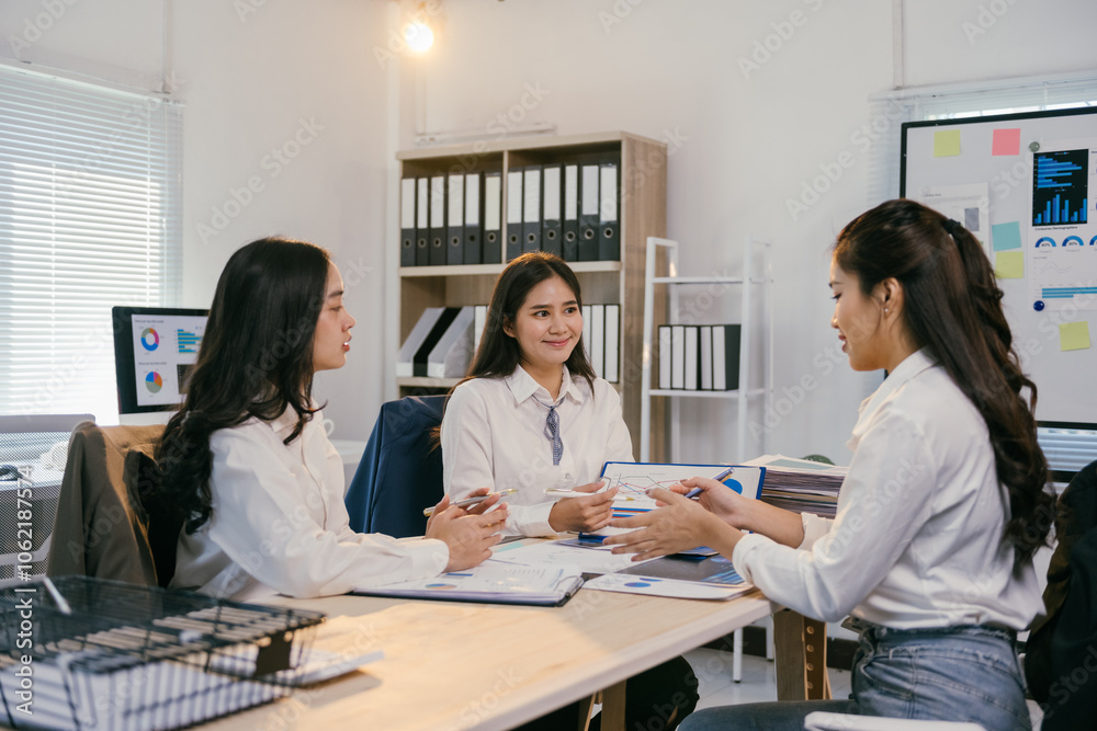 Three asian businesswomen are sitting at a table in a modern office, collaborating on a project and analyzing charts and graphs Three asian businesswomen are sitting at a table in a modern office, collaborating on a project and analyzing charts and graphs