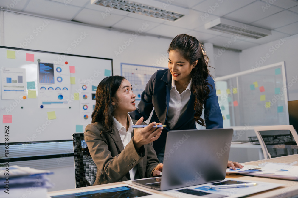 Two asian businesswomen are discussing over financial charts using laptop in a modern office with whiteboard full of sticky notes in the background Two asian businesswomen are discussing over financial charts using laptop in a modern office with whiteboard full of sticky notes in the background