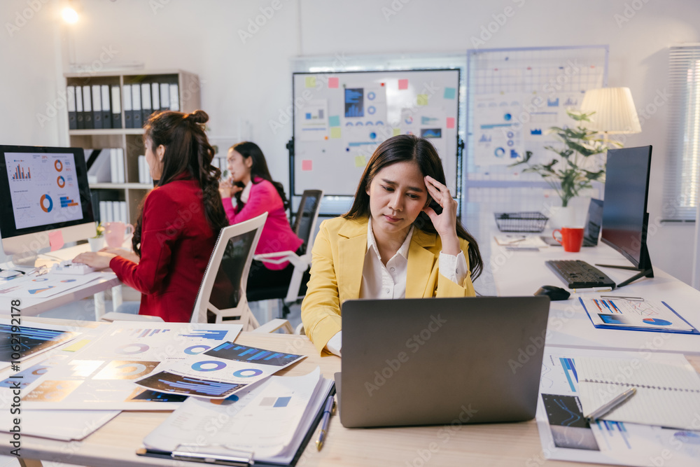 Young professional woman suffering from headache while analyzing financial charts on laptop, showing symptoms of fatigue and burnout in busy office environment with colleagues working in background Young professional woman suffering from headache while analyzing financial charts on laptop, showing symptoms of fatigue and burnout in busy office environment with colleagues working in background