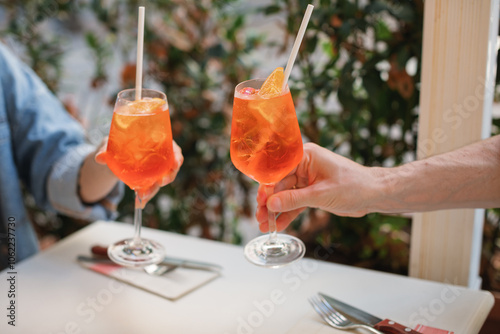 Close-up of a man and woman at an outdoor restaurant clinking glasses with orange spritz cocktails