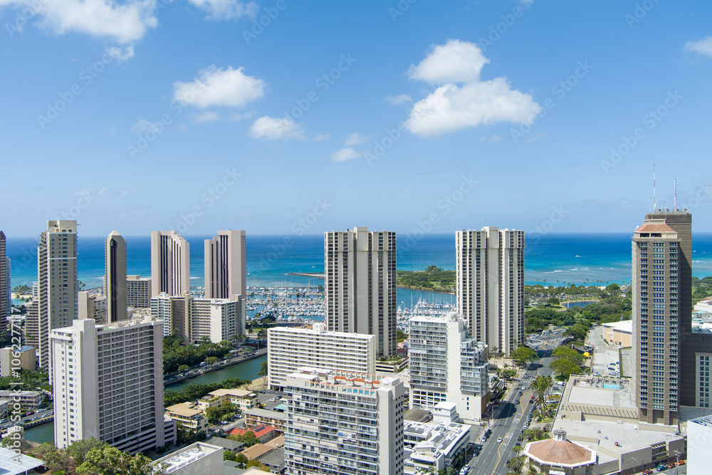 Fototapeta premium aerial shot of the Ala Wai Canal with skyscrapers, hotels and condos, lush green trees and cars driving on the street, boats and yachts in the harbor in Honolulu Hawaii USA