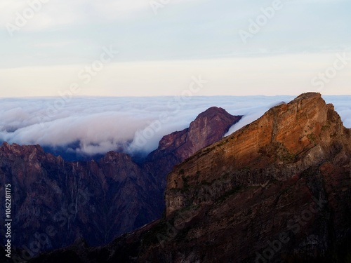 Sharp mountain peaks pierce through a thick flow of clouds as twilight casts warm red hues upon them, creating a dramatic and awe-inspiring natural scene - Pico do Arieiro, Madeira, Portugal.