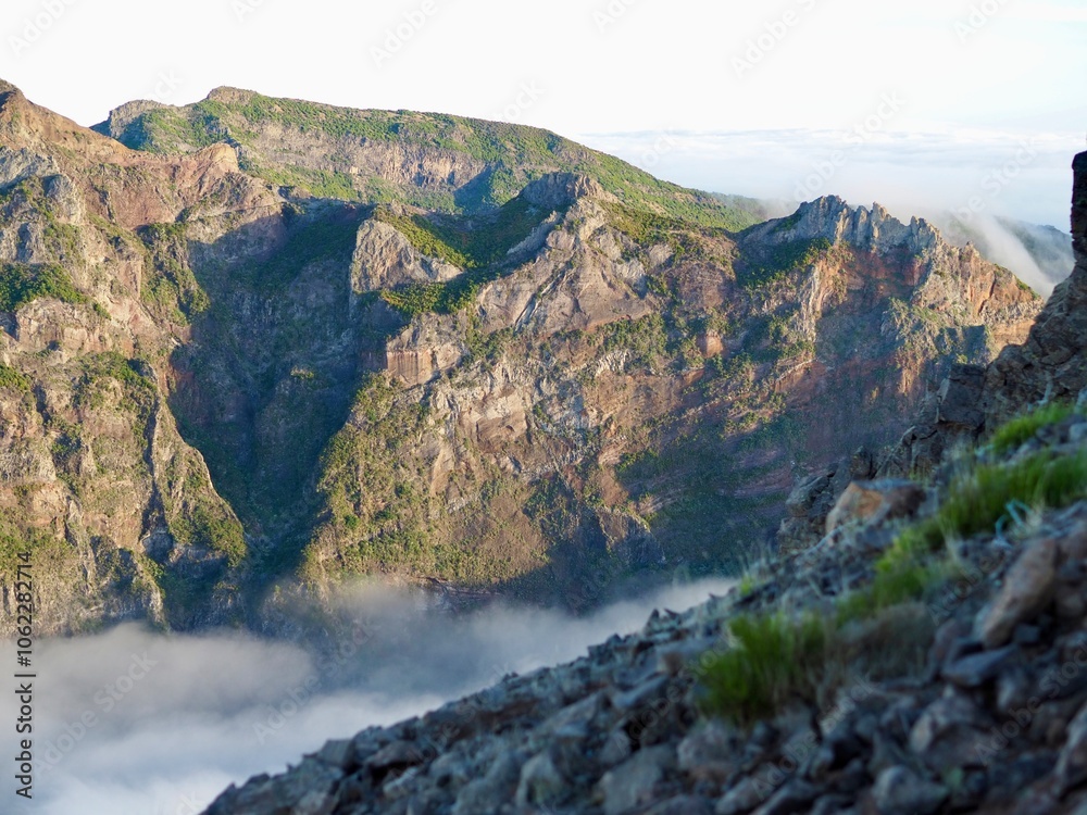 Obraz premium A breathtaking view of rugged mountain cliffs bathed in sunlight, enveloped in dense white fog, and thick lush greenery cascading down the slopes - Pico do Arieiro, Madeira, Portugal.