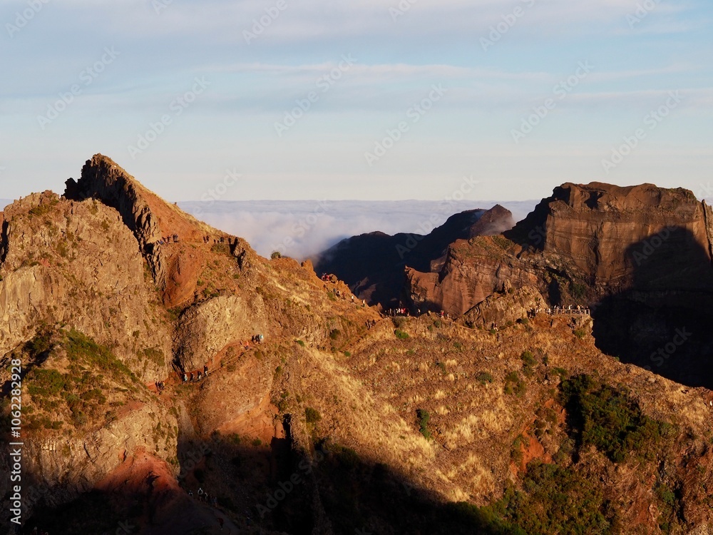 Fototapeta premium An awe-inspiring rocky ridge illuminated by the golden hour light, with a trail of hikers moving along the edge, contrasting with the cloudy horizon beyond - Pico do Arieiro, Madeira, Portugal.