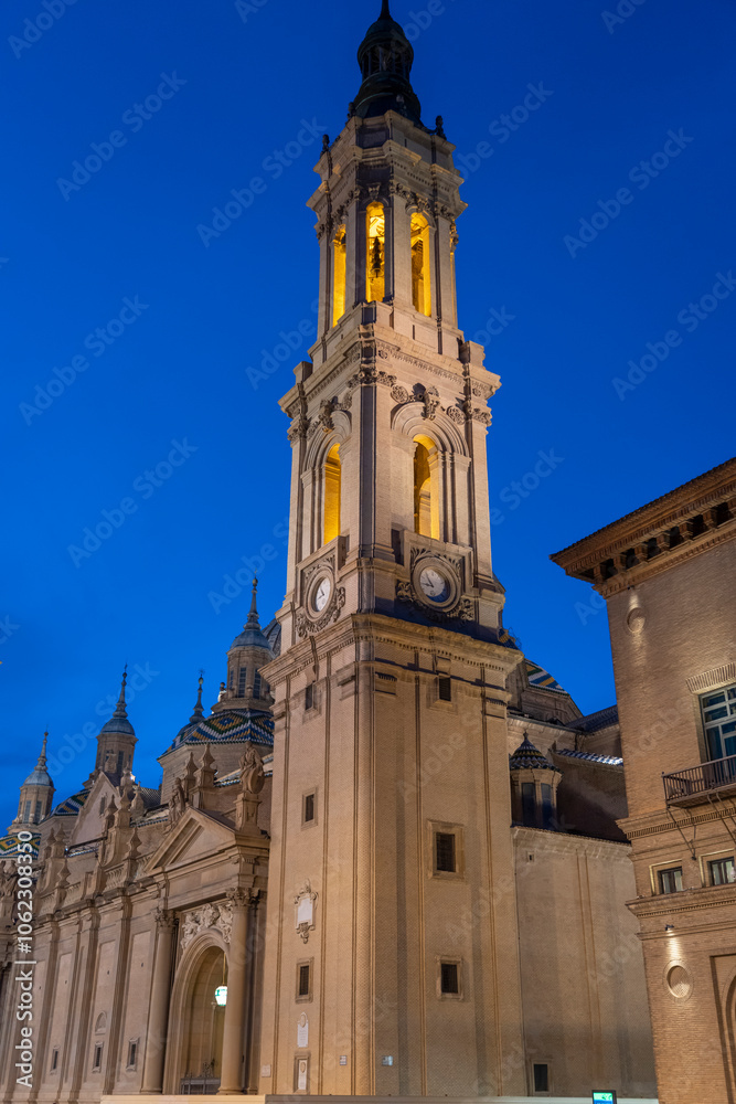 Fototapeta premium Illuminated Basilica del Pilar in Zaragoza, Spain, at dusk, with serene reflections on the Ebro River. Ideal for travel, architecture, and cultural themes.