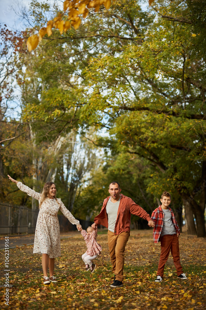 Obraz premium Happy family walking on green grass strewn with fallen leaves in Park in autumn. Parents, two children are walking in park at sunset with holding hands. Family, child, walk through Autumn Park.