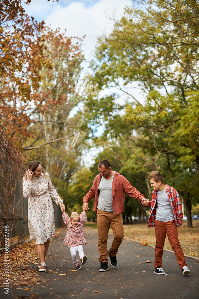 Fototapeta premium Happy family walking on green grass strewn with fallen leaves in Park in autumn. Parents, two children are walking in park at sunset with holding hands. Family, child, walk through Autumn Park.