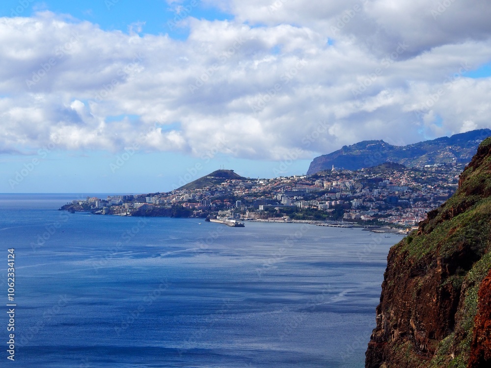 A breathtaking view of a coastal city nestled between the ocean and mountains, with clouds overhead, symbolizing harmony, grandeur, and nature magnificence - Madeira, Portugal. A breathtaking view of a coastal city nestled between the ocean and mountains, with clouds overhead, symbolizing harmony, grandeur, and nature magnificence - Madeira, Portugal.