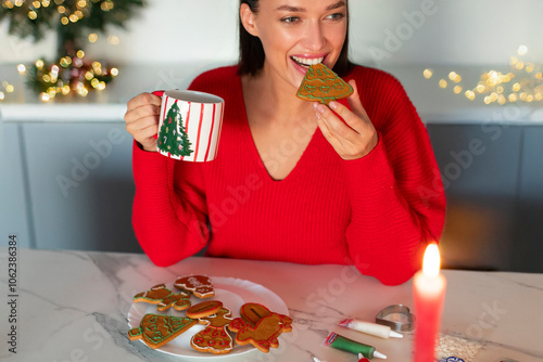 Woman in red sweater celebrating Christmas, eating homemade cookies and drinking cocoa or tea at home in kitchen interior