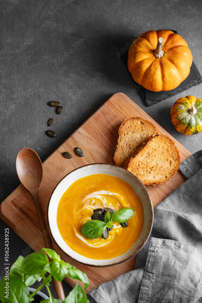 Pumpkin soup with cream, basil and seeds on a wooden board with fried bread, fresh herb and spoon on a dark background.