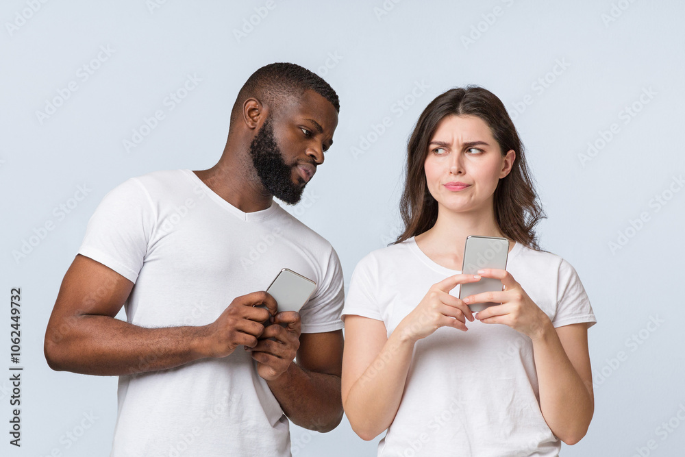 Curious Black Man Spying And Peeking At Smartphone Of His Girlfriend While They Standing Together Over Light Background In Studio, Empty Space Curious Black Man Spying And Peeking At Smartphone Of His Girlfriend While They Standing Together Over Light Background In Studio, Empty Space