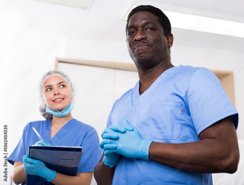 Obraz premium Portrait of confident african american male cosmetologist standing in modern aesthetic medicine office on background with female assistant filling out medical form..