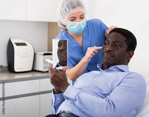 Woman beautician in mask examining face skin of afro american male patient before cosmetology procedure in beauty clinic photo