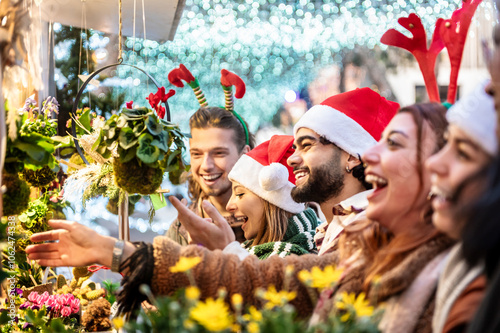 Happy group of friends having fun at Christmas market souvenir shop - Young cheerful tourists wearing santa claus hat enjoying winter holidays outdoors - Traditional culture and holidays concept