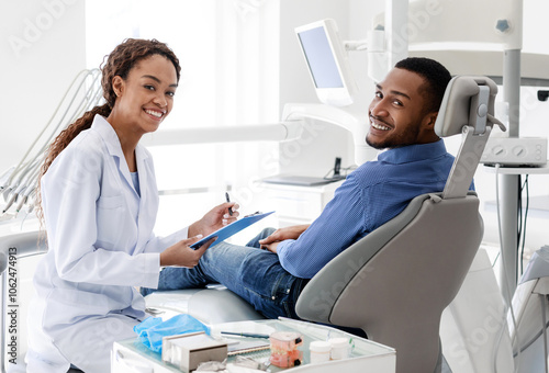 Smiling black female dentist talking to patient and filling medical form in cabinet, free space