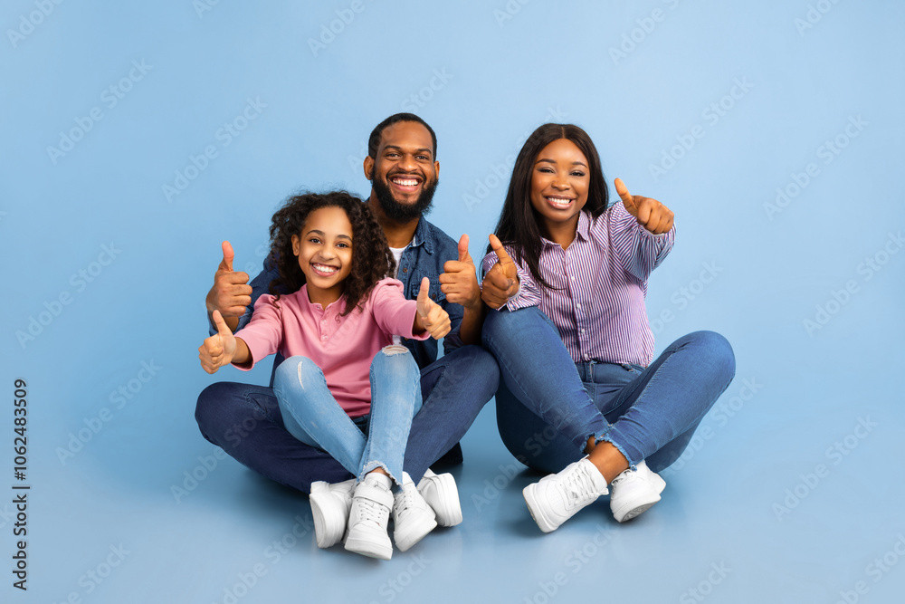 Like Concept. Portrait of smiling black family showing thumbs up gesture with both hands, approving or recommending something good. Mom, dad and daughter sitting on the floor at blue studio Like Concept. Portrait of smiling black family showing thumbs up gesture with both hands, approving or recommending something good. Mom, dad and daughter sitting on the floor at blue studio