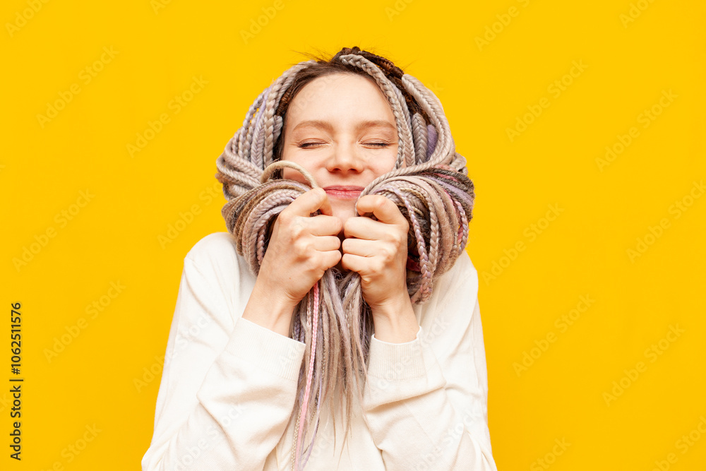 young cheerful girl with gray dreadlocks touches the soft ends of her hair and smiles on a yellow isolated background, woman with an unusual hairstyle and pigtails