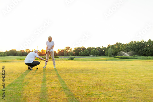 old man and woman in uniform playing golf on golf course at sunset, elderly couple having active outdoor recreation and doing sports, grandpa and grandma holding clubs and pointing where to hit the photo