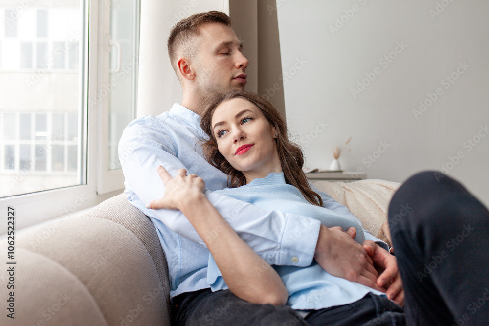 young happy couple in love sitting and hugging on the sofa at home, thoughtful woman lying with boyfriend looking out the window and thinking young happy couple in love sitting and hugging on the sofa at home, thoughtful woman lying with boyfriend looking out the window and thinking