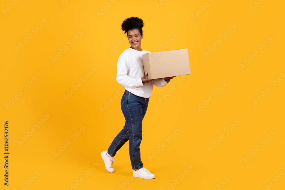 A young woman with curly hair smiles as she walks while holding a cardboard box. The bright yellow backdrop adds a cheerful touch to the scene, reflecting a positive mood. A young woman with curly hair smiles as she walks while holding a cardboard box. The bright yellow backdrop adds a cheerful touch to the scene, reflecting a positive mood.