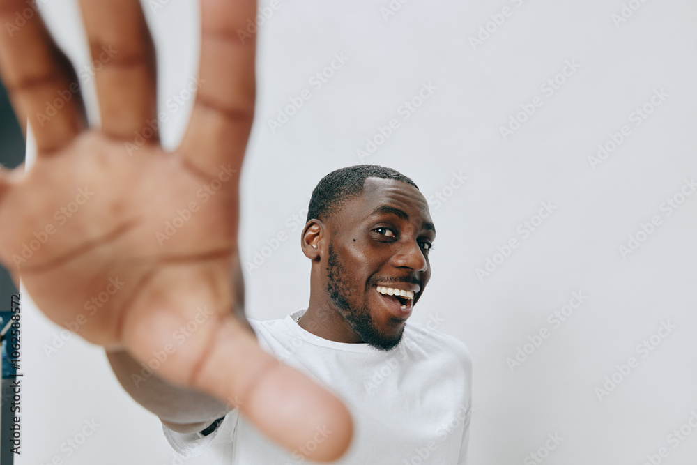 Smiling young man with hand raised, expressing joy, wearing a white shirt against a light backdrop, showcasing confidence and happiness Engaging and cheerful demeanor for lifestyle image Smiling young man with hand raised, expressing joy, wearing a white shirt against a light backdrop, showcasing confidence and happiness Engaging and cheerful demeanor for lifestyle image