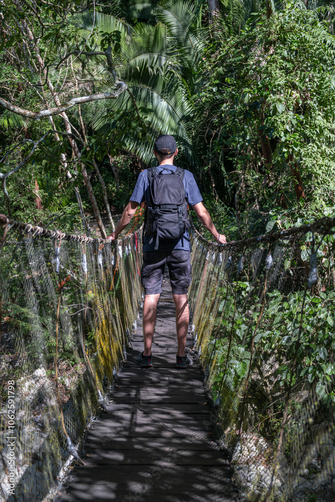 Obraz premium Young man with backpack standing on a suspension bridge surrounded by tropical forest. 