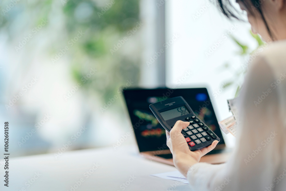 A mother in her home office reviews annual expenses and taxes, with a stock chart displayed on her computer screen, balancing family budgeting and investments. A mother in her home office reviews annual expenses and taxes, with a stock chart displayed on her computer screen, balancing family budgeting and investments.