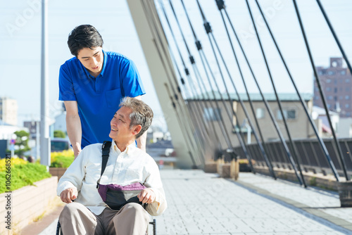 介護、介護士、車椅子、外出、ヘルパー、シニア、高齢者、お年寄り、街、笑顔、都市、社会、ウェルネス、福祉、ケアスタッフ、スタッフ、介助、老後、介護福祉士、橋、道路、歩道、バリアフリー、障害者、障害、ホームヘルパー、散歩、屋外、サービス、サポート、訪問、介護員、自立支援、リハビリ、健康、ヘルスケア、デイサービス、老人ホーム、付き添い、施設、青空、ポジティブ、元気、明るい、リラックス、男性、日本人、歩く