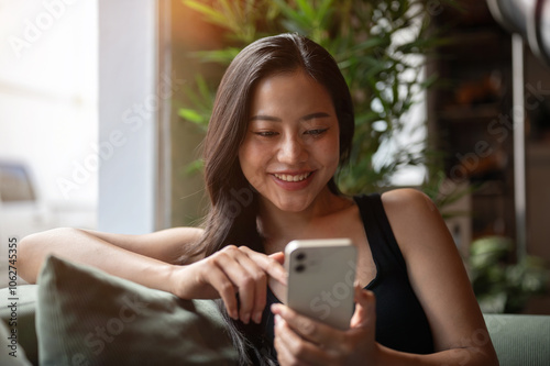 A beautiful Asian woman relaxing on a sofa, smiling as she enjoys chatting on her smartphone.