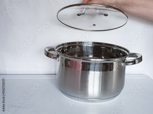 Female hand of housewife opens glass lid of stainless steel saucepan. Kitchen metal utensils stands on table. Background, place for text. photo