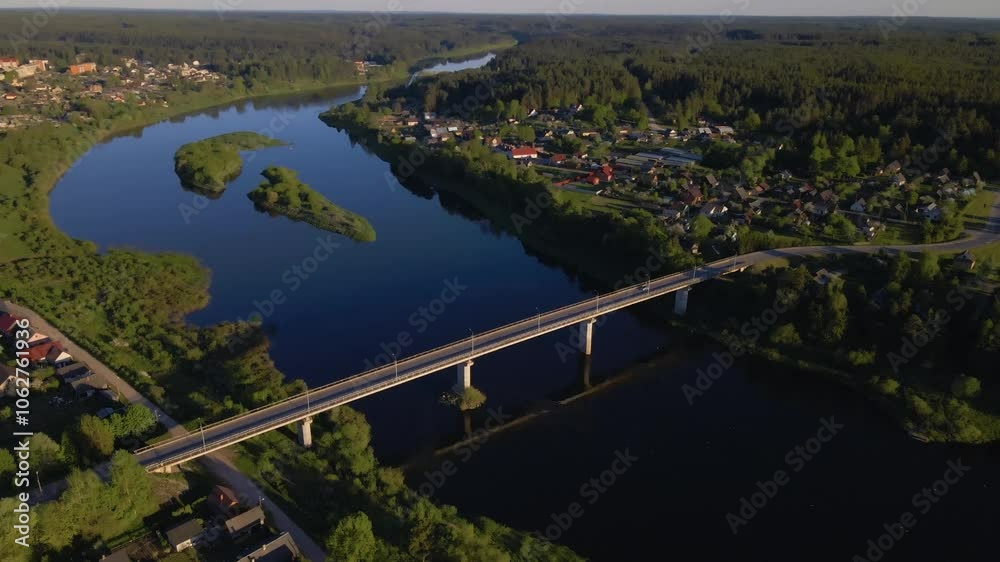 Drone tracks a car driving across a bridge over a river slowly circling to reveal a picturesque town and vibrant nature. The scene captures reflections on the water during golden hour. Drone tracks a car driving across a bridge over a river slowly circling to reveal a picturesque town and vibrant nature. The scene captures reflections on the water during golden hour.