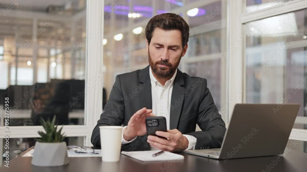 Professional financial advisor, executive leader, manager, businessman sits at his desk in office, using his mobile phone and laptop. Office worker is focused on his work and appears to be productive. Professional financial advisor, executive leader, manager, businessman sits at his desk in office, using his mobile phone and laptop. Office worker is focused on his work and appears to be productive.