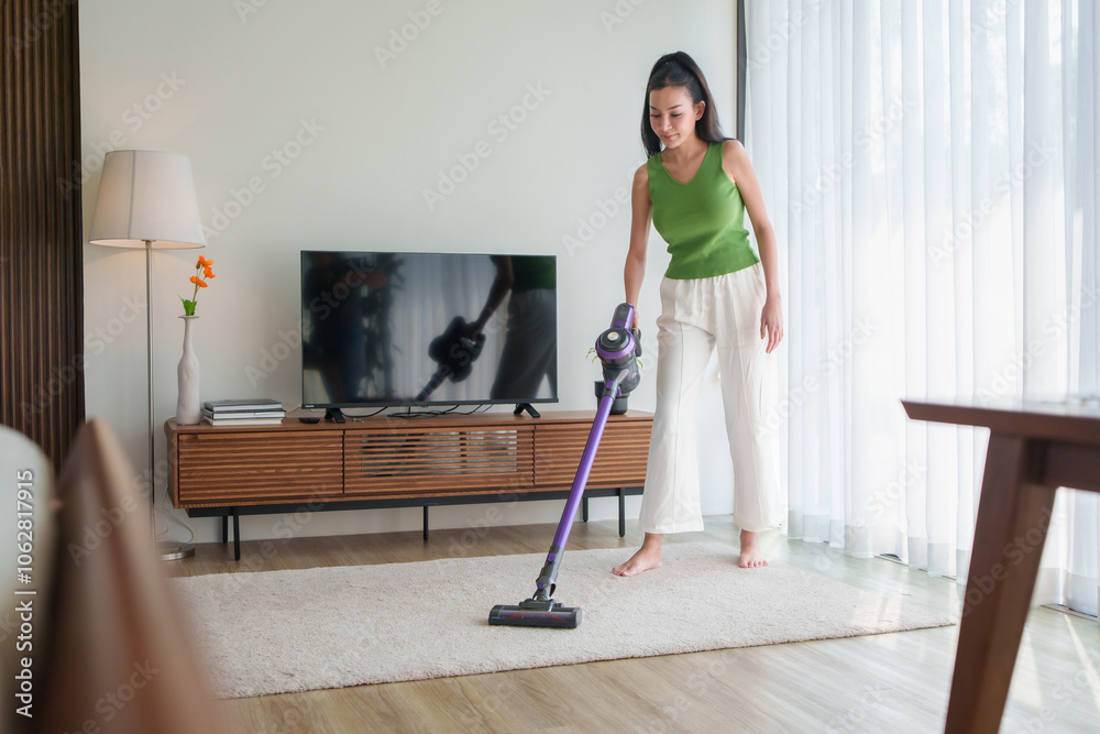 Young Asian Woman vacuuming a living room rug with a cordless vacuum cleaner for housekeeping housework and chores in a modern home. Young Asian Woman vacuuming a living room rug with a cordless vacuum cleaner for housekeeping housework and chores in a modern home.