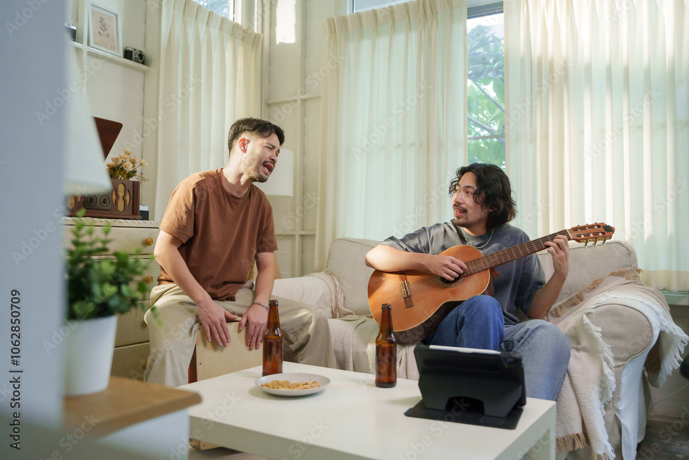 Asian playing guitar while his friend plays a cajon, both singing and enjoying a musical jam session in a living room. Friendship concept. Asian playing guitar while his friend plays a cajon, both singing and enjoying a musical jam session in a living room. Friendship concept.
