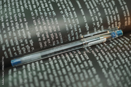 A beautiful, unique styled book is wide opened with black colored shiny pages and white colored Bangla writings, also with a blue colored gel pen is placed in the middle of the book.