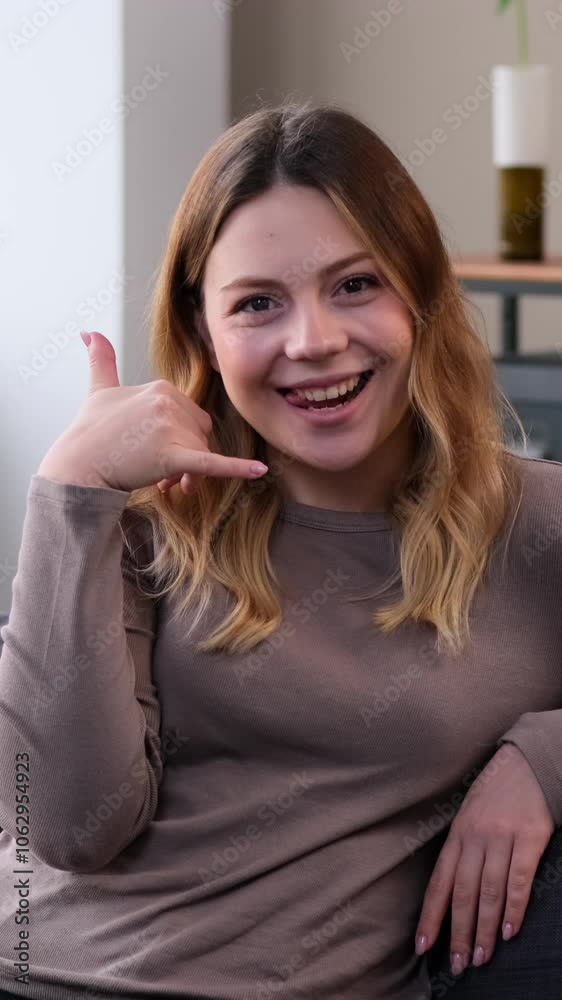 Welcoming young adult Caucasian woman smiling and showing call me gesture at home. Concept of a communication and invitation. Vertical video.