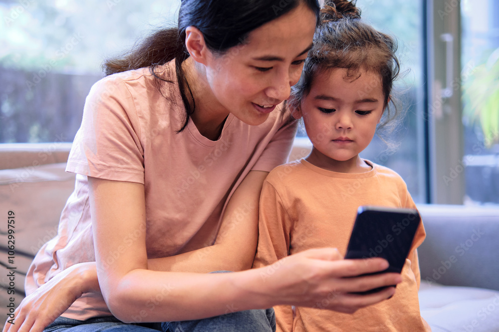 Smiling woman sharing mobile phone with daughter at home Smiling woman sharing mobile phone with daughter at home