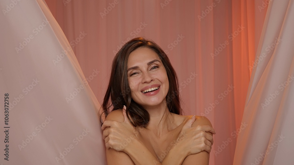 Woman with makeup and hairstyle looks at camera smiles, touches neck and chest skin area, flying pink fabrics studio background. Beauty and wellness concept. Woman with makeup and hairstyle looks at camera smiles, touches neck and chest skin area, flying pink fabrics studio background. Beauty and wellness concept.