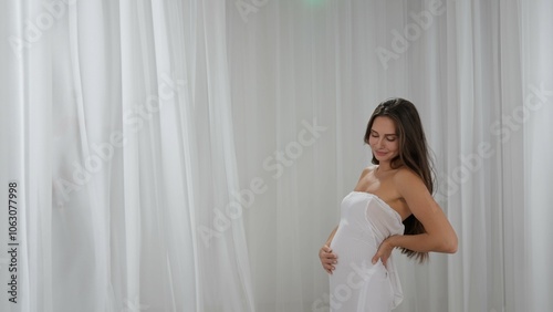 Woman in dress with natural makeup and hairstyle looks at camera, smiling and touching her pregnant belly, flying white fabrics studio background. Beauty and wellness concept.