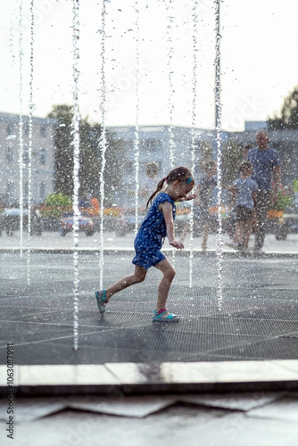 Little girl running over fountain. Hot summer weather in city. Child  playing near fountains in hot weather. Cute girl running between water jets of fountains.