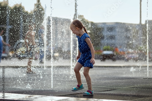 Little girl running over fountain. Hot summer weather in city. Child  playing near fountains in hot weather. Cute girl laughing between water jets of fountains.