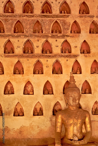 A big golden, gilded buddha statue, sculpture in front of a wall filled with many miniature buddha figurines in the cloister wall of Wat Si Saket, a buddhist temple in Vientiane, Laos photo